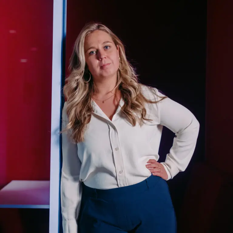 Een ZZP-vrouw met lang blond haar, een witte blouse en een blauwe broek staat binnen met een hand op haar heup en een neutrale uitdrukking, leunend tegen een lichte muur tegen een donkerrode achtergrond.