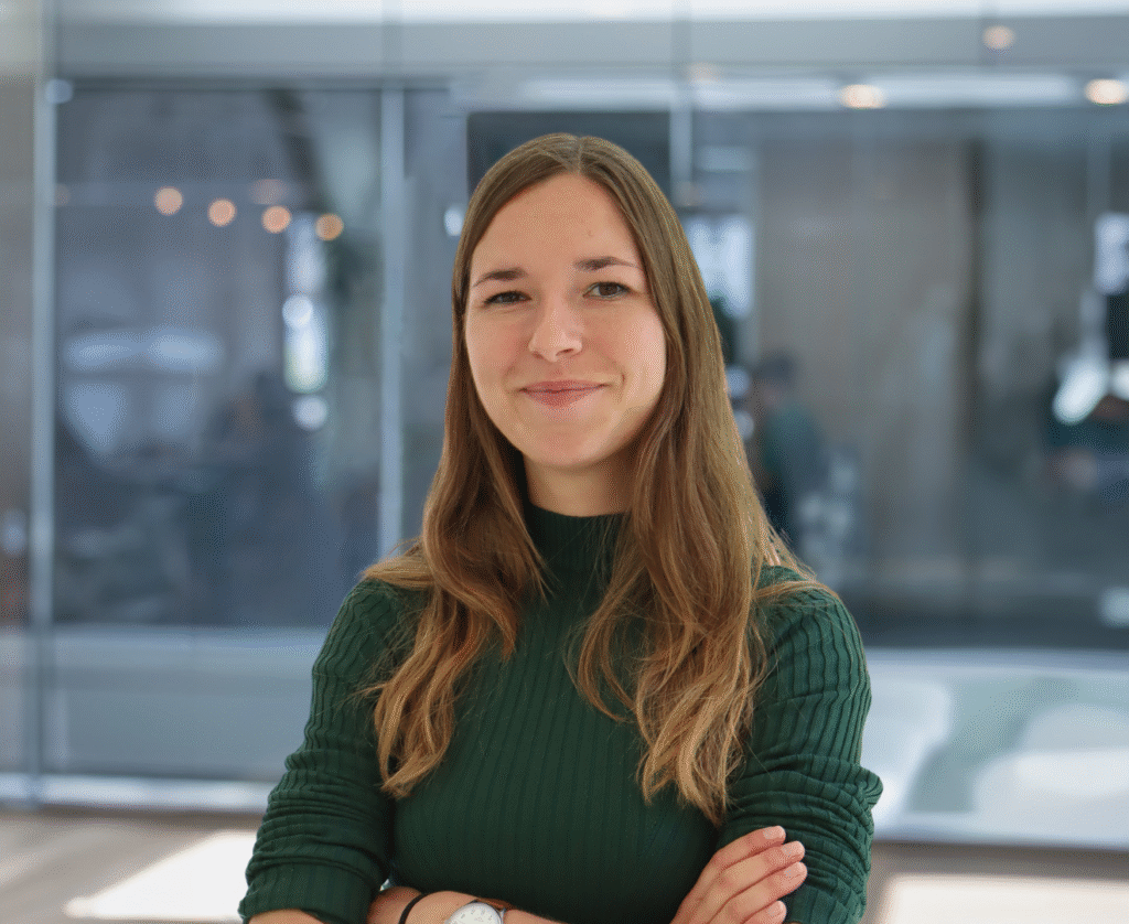 Een vrouw met lang bruin haar, die een groene geribbelde trui draagt, staat binnen met haar armen over elkaar, glimlacht lichtjes en straalt het zelfvertrouwen uit van een ervaren beleidsadviseur. De achtergrond toont een glazen wand en een vage kantooromgeving.
