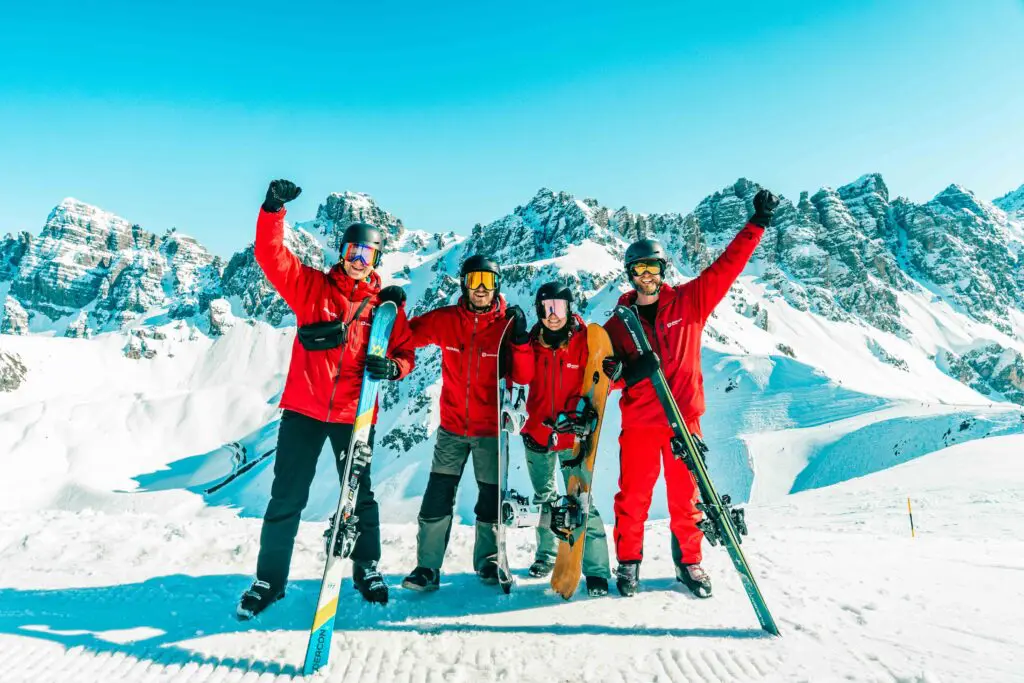 Vier mensen met rode jassen en winteruitrusting staan op een besneeuwde berghelling met ski's en een snowboard in de hand. Ze glimlachen en steken hun armen feestelijk omhoog met besneeuwde bergtoppen op de achtergrond.