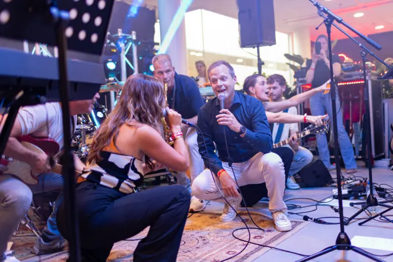 Een levendige band treedt op op het podium; een vrouw en een man vooraan zingen in microfoons, omringd door muzikanten die instrumenten bespelen, met kleurrijke podiumlichten en een juichende menigte op de achtergrond.