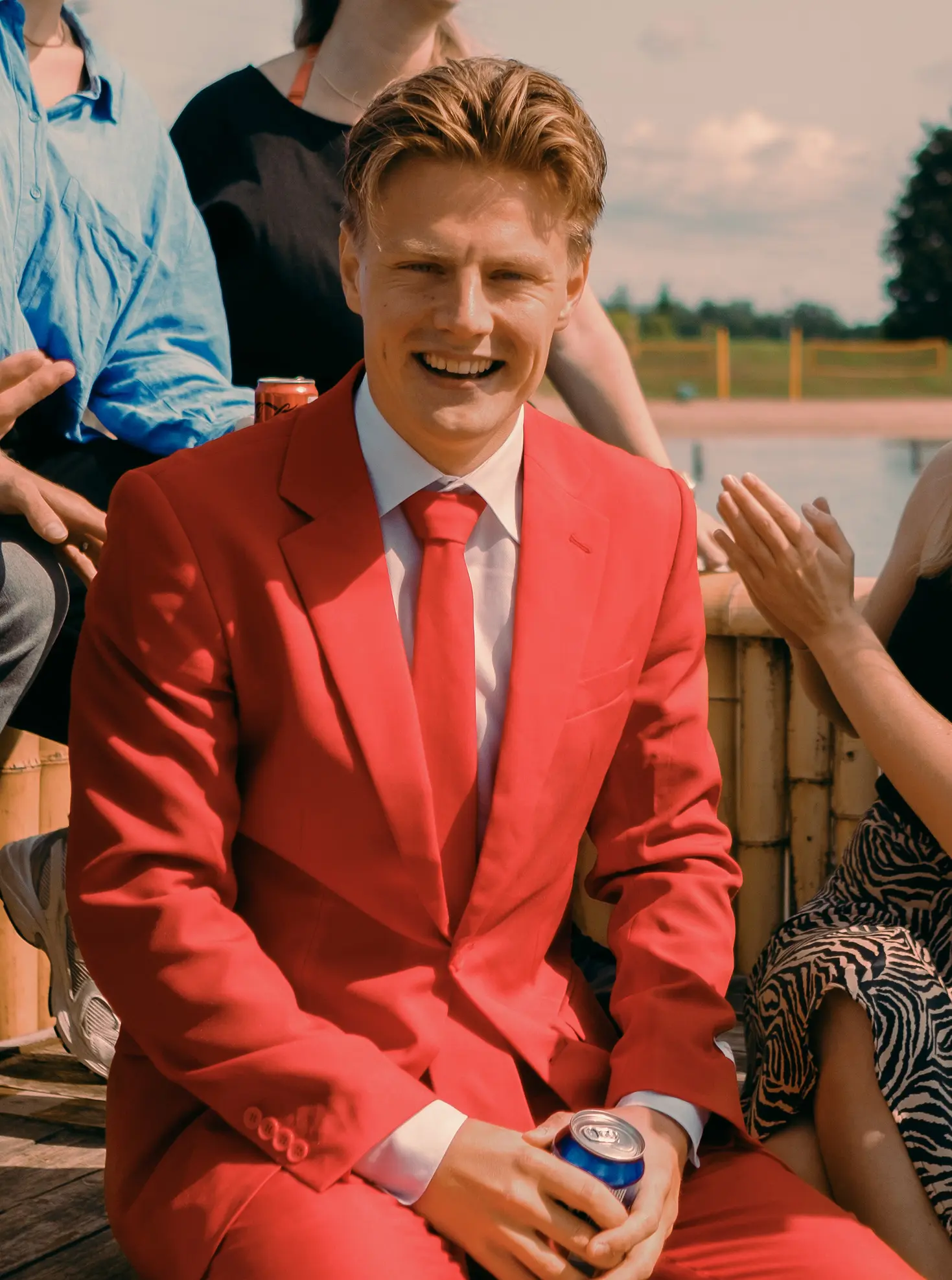 Man in een rood pak met een drankje, zittend buiten bij zonnig weer met anderen op de achtergrond.