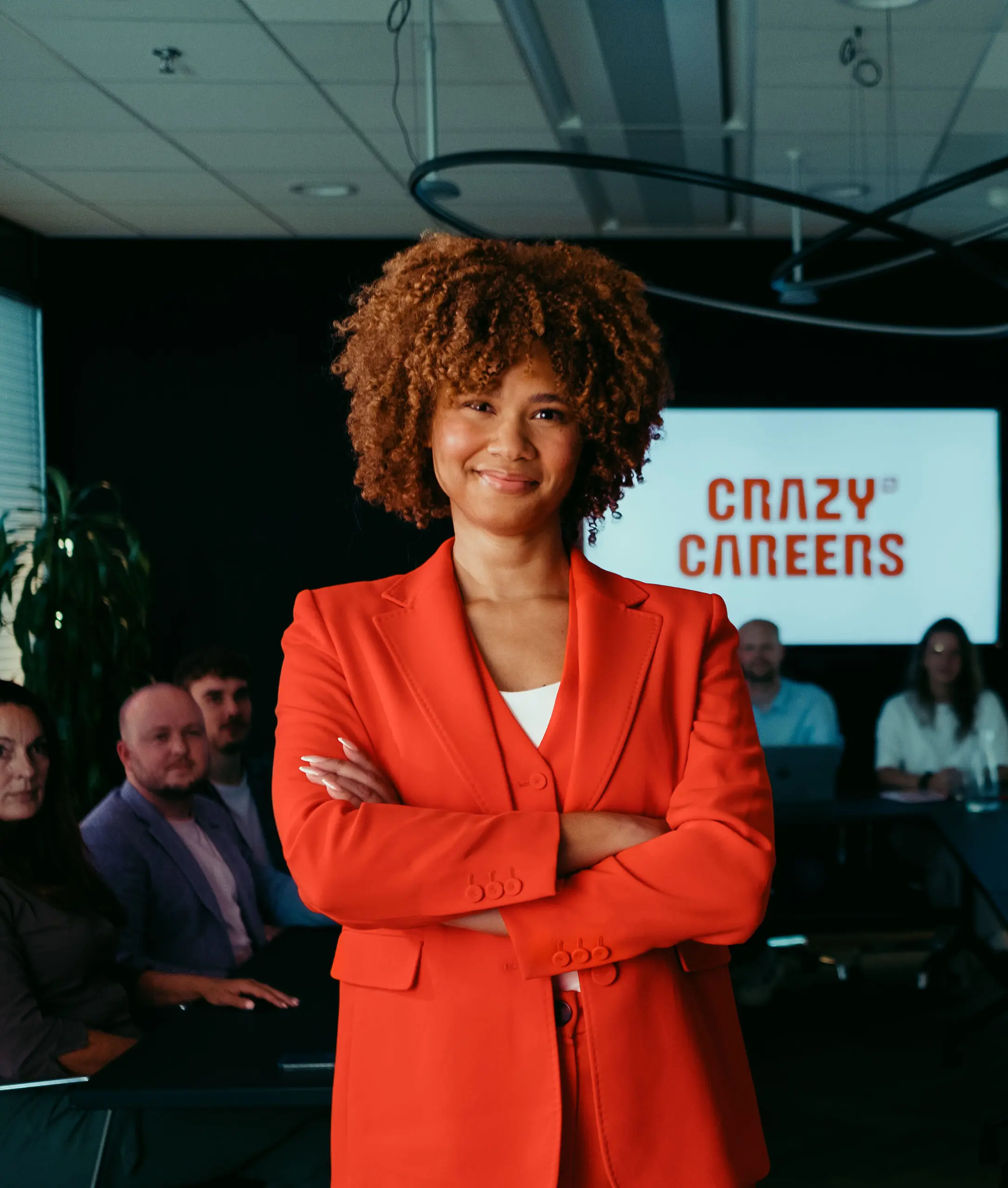 Een vrouw in rood pak staat voor een scherm met "Crazy Careers", met mensen op de achtergrond.