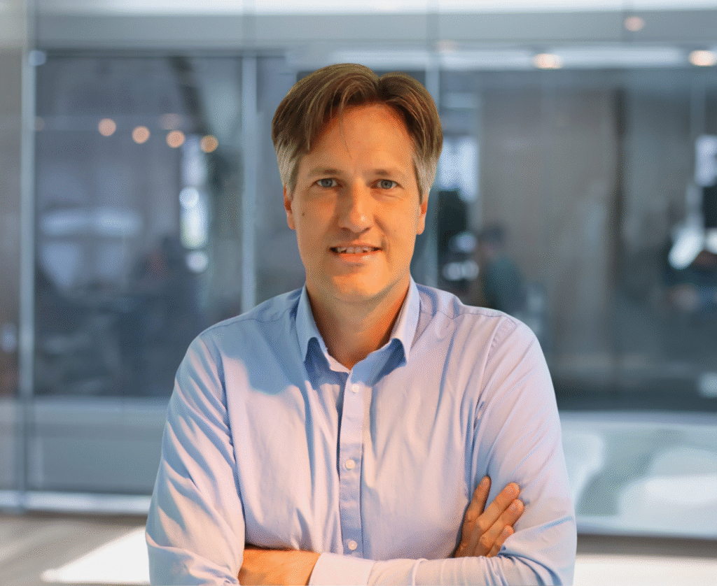 Een man in een blauwe overhemd staat met gekruiste armen in een moderne kantooromgeving.